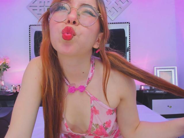 KattyHards with red pigtails, wearing a pink floral halter top, pouting her red lips, wearing round glasses, in a bedroom with pink and purple lighting.