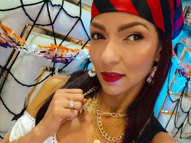 HannaPretty has brown skin, red lipstick, and black-red striped pirate hat, with long red hair, wearing gold chains, earrings, and a black-white top, showing her tits.