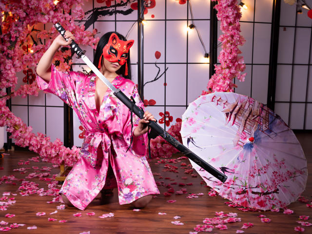 AnyaJay in pink floral kimono, red fox mask, holding katana, kneeling with cherry blossom petals, white umbrella with blood splatter, wooden floor, string lights.