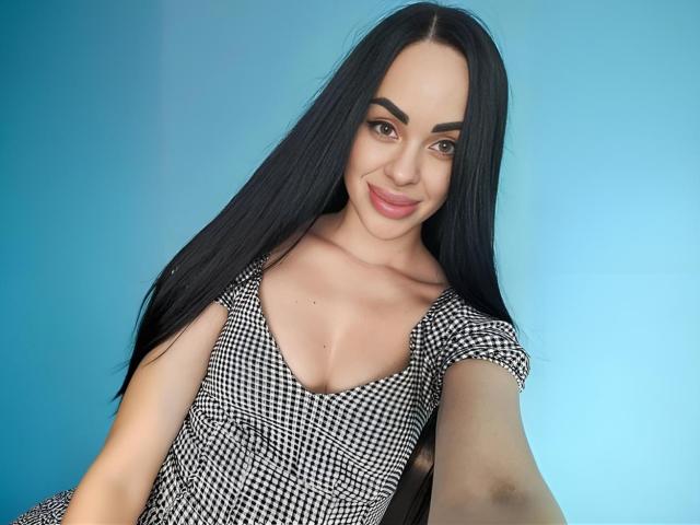 Stefaniya, met lang zwart haar, draagt een zwart-wit geruit jurkje en glimlacht naar de camera. Ze laat op een selfie haar kleine borsten zien, met een blauwe achtergrond..