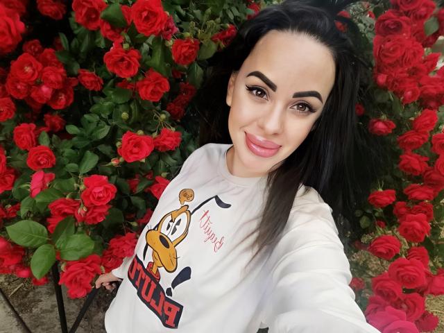 Stefaniya, a woman with long black hair and dark eyebrows, takes a selfie. She wears a white sweatshirt with a cartoon duck and "UNIDUCK" text. She has pink lipstick and a slight smile. She's in front of a dense background of red roses. The image is bright with vivid colors, focusing on her face and the floral backdrop. The composition is tight, emphasizing Stefaniya's upper body and the roses behind her.