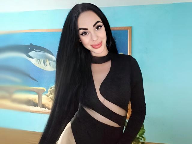 Stefaniya with long black hair, wearing a black, cut-out, long-sleeve top, showing her small tits and a smile, in a room with a blue wall and shark painting.