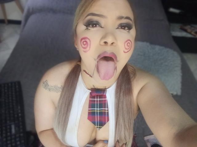 TatianaRossi with blonde pigtails, white halter top, plaid necktie, and pink swirls on cheeks, tongue out, tattoos, and large tits, in a selfie with a playful expression.