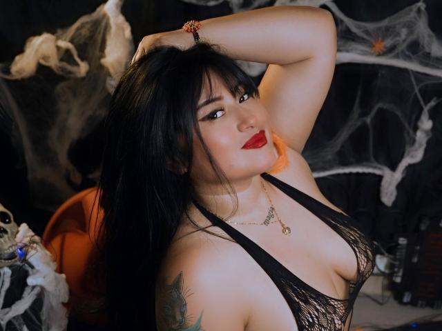 KimoraKim com lingerie de renda preta, batom vermelho e cabelo preto longo, com a mão no cabelo, mostrando os seios, uma tatuagem no braço e um colar..