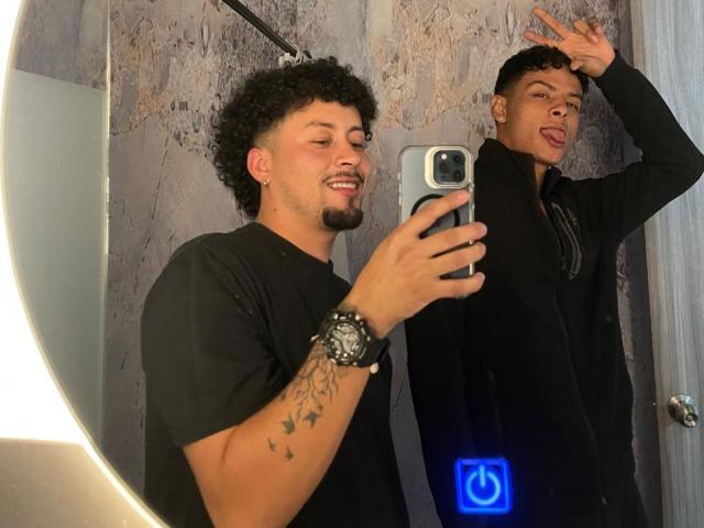 Twee mannen met krullend, kort zwart haar, die zwarte shirts dragen, maken een selfie in een badkamer. De ene heeft zijn tong uitgestoken, de andere glimlacht, en draagt een horloge om zijn pols..