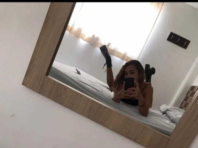 A mirror reflection shows JessiBlonde lying on a bed, holding a phone with both hands. She has a light brown skin tone, long dark blonde hair, and is wearing black high-heeled shoes with her feet up. She's in a white room with a window covered by beige curtains. The bed has a gray sheet. The mirror has a wooden frame. The photo is casual and slightly tilted.