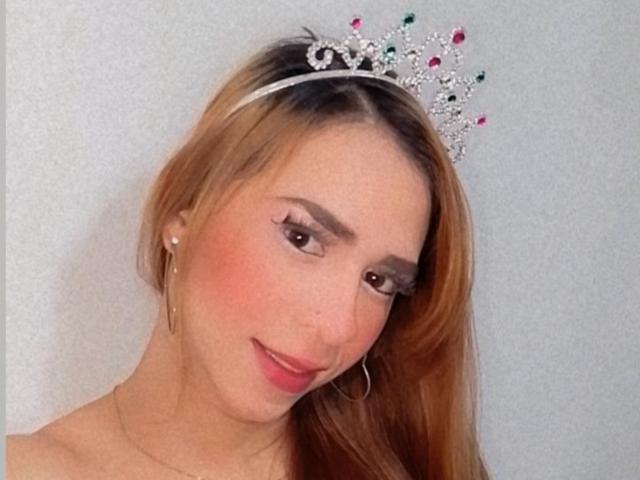 JessiBlonde wears a sparkly silver tiara with pink and green jewels, set against a plain white background. She has long, straight blonde hair, and her face is slightly tilted to the right with a subtle smile. Her skin is fair, and she has pink blush on her cheeks. She's wearing small pearl earrings and a thin gold necklace. Her makeup includes dark eyeliner and long eyelashes. The image is a close-up, focusing on her upper body and face.