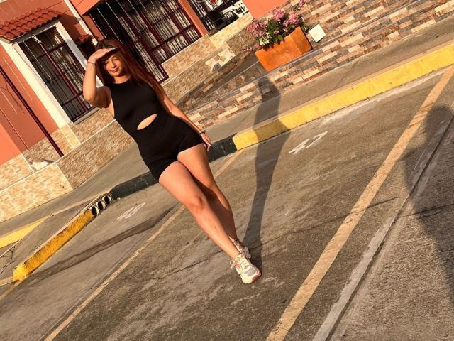 AbbyParkerr in a black cutout dress and shorts, standing in a sunlit parking lot, hand on head, white sneakers, yellow curb, brick wall, orange building, pink flowers.