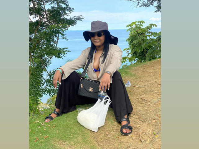 MelanyScoth is squatting with her tits slightly visible in a blue bra, wearing a gray bucket hat, black sunglasses, and black pants, holding a bag and a water bottle.
