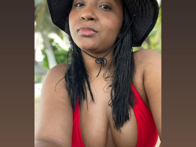 MelanyScoth with wet black hair, wearing a black hat and red bikini top, showing her big, juicy tits. She has a small nose ring and a subtle smile.