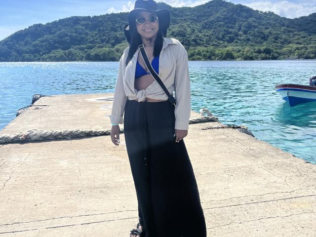 MelanyScoth stands on a concrete dock, wearing a black hat, white shirt tied at the front, blue bikini top, black high-waisted skirt, and sunglasses, with a boat in the background.