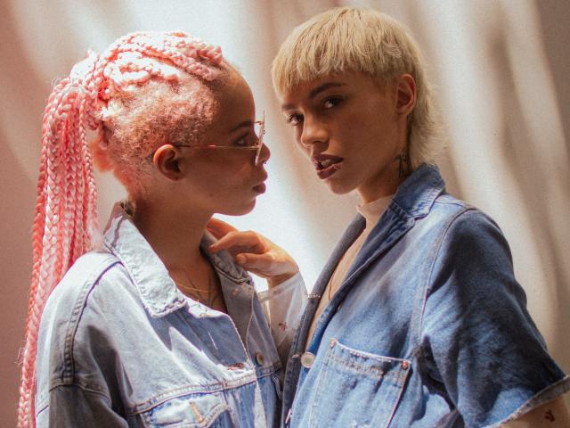 GravesYStarveil with pink braided hair and glasses faces GravesYStarveil with blonde bob, wearing denim jackets. The pink-haired GravesYStarveil has a gold necklace, while the blonde GravesYStarveil has a white shirt underneath. Light from the side highlights their faces, creating a contrast between their hair colors. Both have intense, focused expressions, with the blonde GravesYStarveil's hand resting on the pink-haired GravesYStarveil's shoulder. Soft, warm lighting.