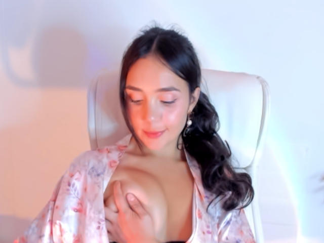LinsayColemann with dark hair in a ponytail, wearing a floral robe, covering her tits with one hand, sitting on a white chair, smiling softly.