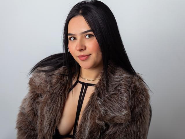 MiaAylen with long black hair, wearing a fur coat, exposing her tits in a black strappy top, smiling at the camera. Soft, natural makeup, light skin.