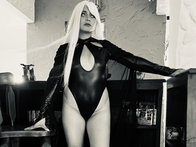EliBaker with long white hair, wearing a black leather cutout bodysuit and long gloves, standing in a room, leaning on a table, showing her tits and ass.