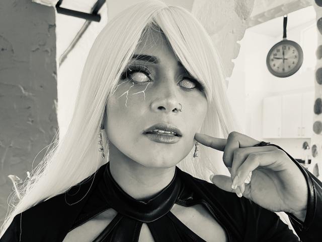 EliBaker with white hair, glowing eyes, and makeup cracks, pointing at nose with finger, wearing black dress with cutouts, ear earrings, and nose ring. Monochrome.