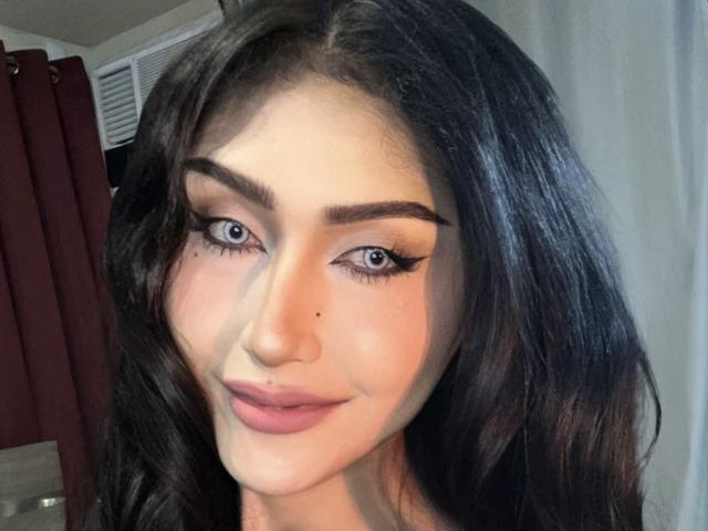 TheaLux has dark wavy hair, blue eyes, and a small beauty mark on her right cheek. She's wearing bold eyeliner and pink lipstick, smiling softly.