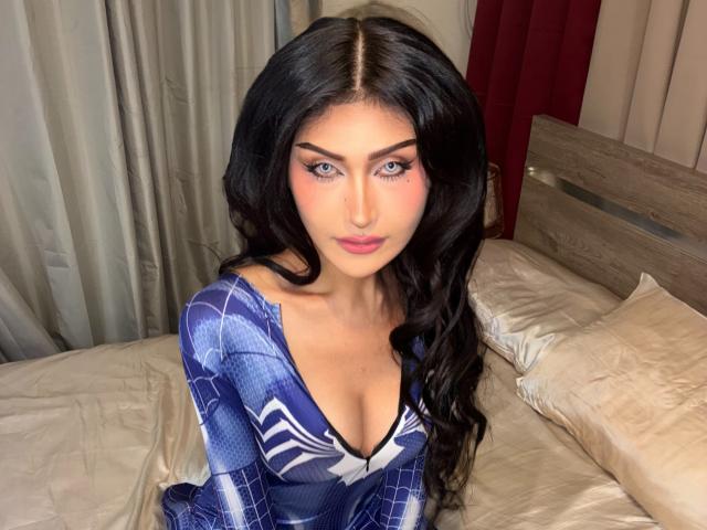 TheaLux with long black hair, blue eyes, and pink lipstick, wearing a blue, patterned dress with cleavage, sitting on a bed with beige pillows and a wooden headboard.