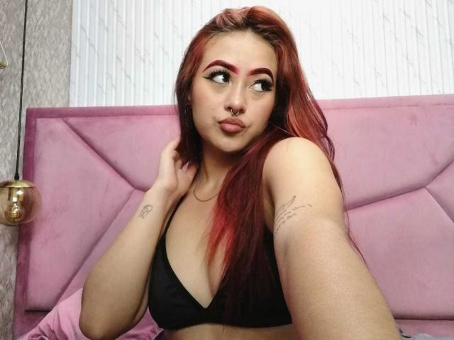 Ein rothaariges Mädchen mit schwarzer BH, Nasenring und Tattoos, das zur Seite blickt, mit voller Lippe, die Hand im Haar, auf einem rosa Bett. Brüste, süß, sexy..