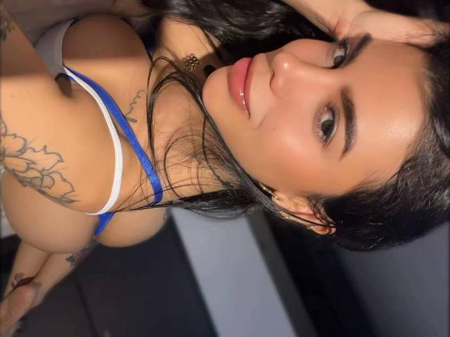 NikolRyder, avec ses cheveux noirs, sa peau bronzée et ses tatouages, porte un soutien-gorge bleu et blanc, regarde l'appareil photo, est légèrement penchée en avant, et montre ses seins et ses fesses..
