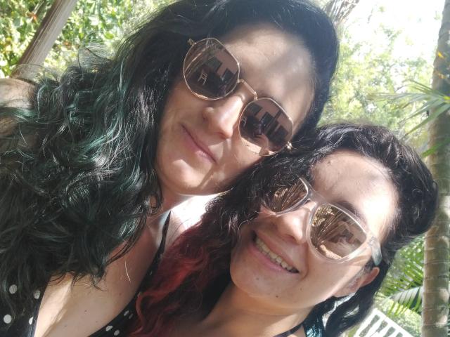 Close-up photo of LunaVenus with curly black hair and LunaVenus with wavy black and red hair. Both wear reflective aviator sunglasses, smiling at the camera. LunaVenus with curly hair is on the left, slightly taller, with a light smile. LunaVenus with wavy hair is on the right, smiling broadly. Bright outdoor light filters through green foliage in the background. Natural textures of hair contrast with the smooth, reflective surfaces of sunglasses. LunaVenus on the right has a small mole on her cheek. Sunlight highlights their faces.