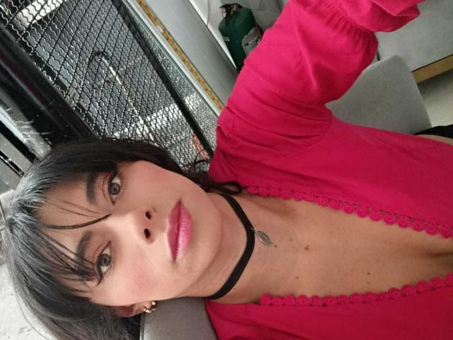 AnnaMooree con un collare nero, labbra rosa e una maglietta rosa, che mostra il seno in un selfie, una maglietta rosa con bordi in pizzo, capelli neri e occhi grigi, in interni..