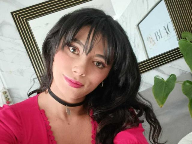 AnnaMooree ha i capelli neri, il rossetto rosa e indossa una collana nera con un piccolo ciondolo. Indossa una maglietta rosa e guarda la fotocamera con un leggero sorriso..