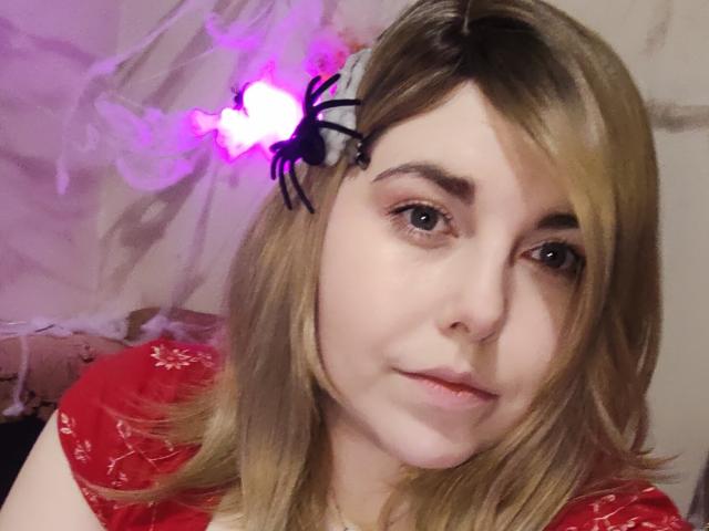 WhitteLotus has blonde hair, a black spider hairclip, and wears a red shirt. She's looking at the camera with a slight smile, showing her ass in the background.