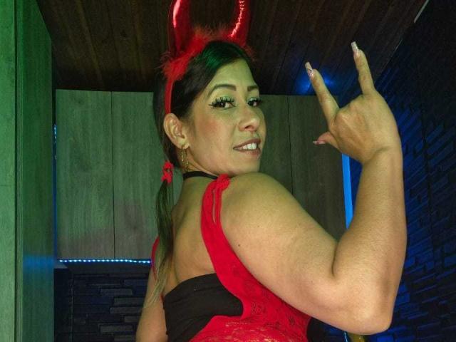 LaurenHills with red devil horns, wearing a red lace top, black strap, and red headband, smiling, showing off her ass with her finger up.
