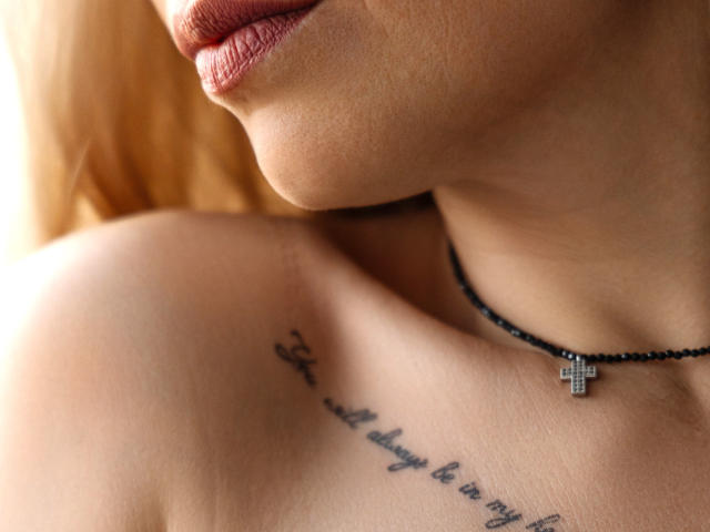 SweetLadyXX's upper chest and lips, showing a tattoo below her tits and a black necklace with a cross. Her skin is light, and her lips are slightly parted.