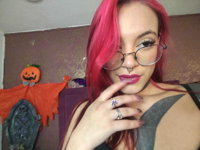 DahianaKlimt with pink hair, glasses, nose ring, and purple lipstick, touching her lips with a finger, showing a black tank top and a tattoo. Halloween pumpkin decoration in background.