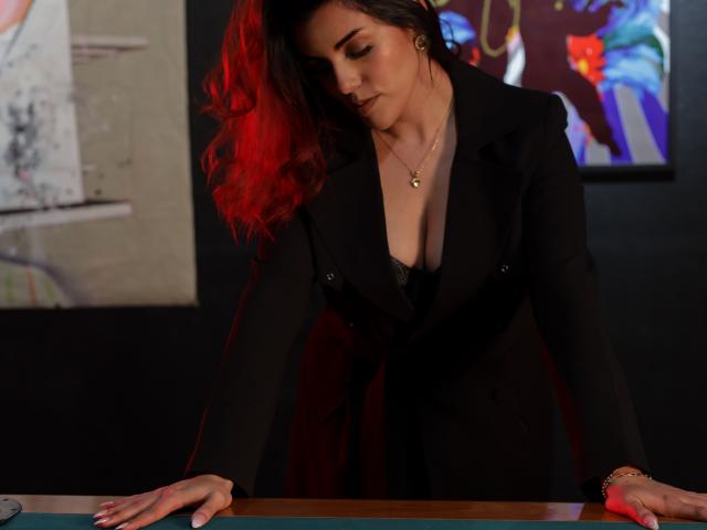 ZoeeMar, avec ses cheveux roux, portant un blazer noir, penchée au-dessus d'une table de billard, montrant ses seins volumineux, un soutien-gorge en dentelle noire, un collier en or et un bracelet, dans une pièce faiblement éclairée..