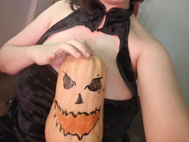 GoddessSophie69 holding a carved pumpkin with a Jack-o'-lantern face, wearing a black, sleeveless, velvety dress, exposing her tits, and her hand is on her chest.