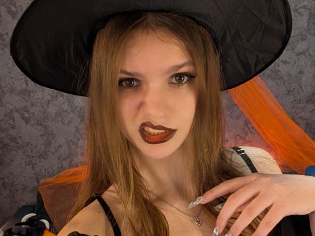 PussyOnLuck with long brown hair, black witch hat, and dark red lipstick, showing off white nails and a necklace, has a seductive, playful expression.