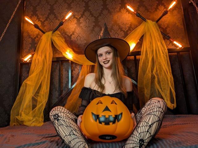 PussyOnLuck with long brown hair, witch hat, black off-shoulder top, fishnet legs, sitting on bed, holding orange pumpkin with jack-o'-lantern face, surrounded by yellow drapes and orange lights.