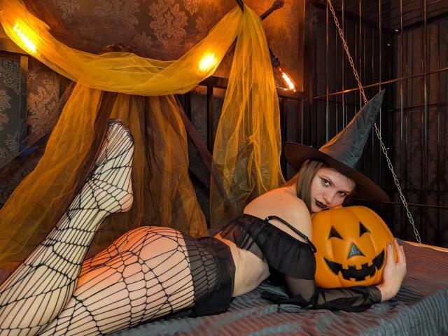 PussyOnLuck lying on bed, wearing black mesh lingerie and witch hat, holding pumpkin with evil grin, legs up, fishnet stockings, yellow and black draped background, dark room.