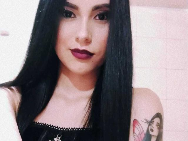 AmberLenox with long black hair, dark lipstick, and a tattoo of a woman with a red flower on her right shoulder, wearing a black top.