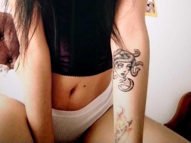 AmberLenox with dark hair, black top, white panties, showing tattoo of a woman's face on right arm and flame tattoo below. Her ass is slightly visible.