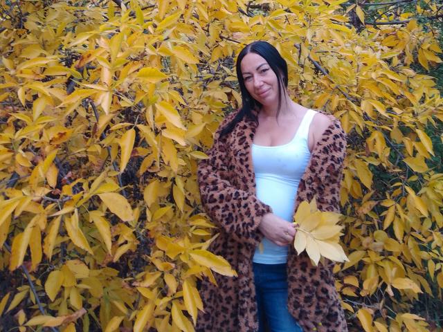 MirandaHott6[9] stands in front of a dense bush of yellow autumn leaves. She has dark hair and a slight smile. She wears a white tank top, blue jeans, and a leopard-print coat. Her right hand holds a yellow leaf. The bright yellow leaves contrast with her dark hair and coat. The image uses natural light. The texture of the leopard-print coat is smooth and shiny. The overall composition is balanced and vibrant.
