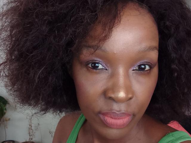 PrettyAndiy, avec ses cheveux noirs et bouclés, sa peau brune, son maquillage pour les yeux violet, son rouge à lèvres rose, son débardeur vert, et une expression sérieuse, regardant directement l'appareil photo..