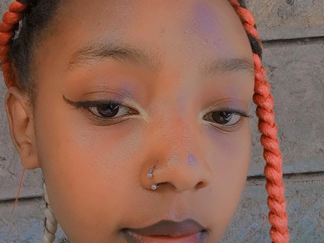 SkyNikey's face with orange braided hair, dark eyeliner, and a nose piercing with two small jewels, against a grey brick wall. She's smiling subtly.