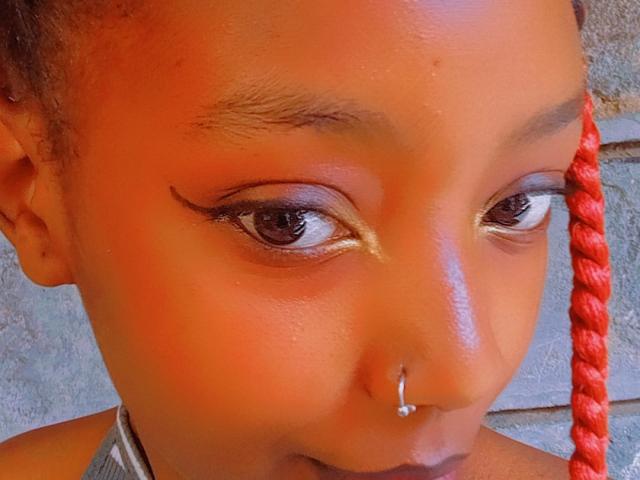 SkyNikey's face, featuring dark brown eyes with gold eyeshadow, a red braid, a silver nose ring, and a shiny skin texture. Her eyebrows are slightly raised.