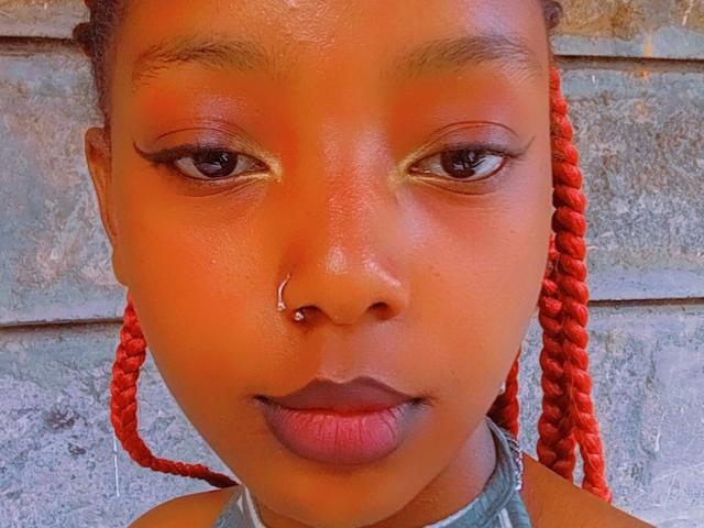 SkyNikey has dark red braided hair, a nose ring, and brown eyes with subtle eyeliner. She's wearing a gray halter top and has a smooth, brown-skinned face.