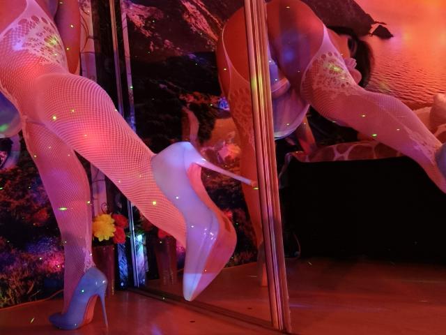 AmberSunshine in fishnet lingerie, blue high heels, and white stockings, reflected in a mirror, showing her ass and reflection, with colorful lights and a flower on the floor.