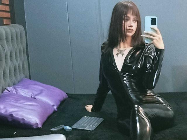 CamiMonet with long brown hair, wearing a shiny black latex bodysuit, sitting on a black carpet, taking a mirror selfie, showing her tits and a purple pillow beside her.
