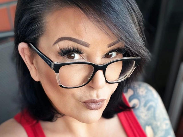 FireFox80-hot with black glasses, dark brown eyes, thick eyebrows, and black shoulder-length hair, wearing a red top, showing a tattoo on her right arm, and a slight smirk.