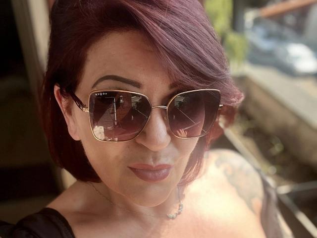 FireFox80-hot with short red hair, wearing dark sunglasses and a black top, showing tattoos on her right arm, and a subtle smile, with sunlight reflecting in her glasses.