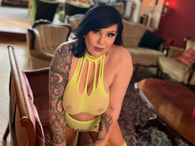 FireFox80-hot with black hair, tattoos on her right arm, wearing a yellow mesh halter that exposes her tits and nipples, kneeling in a cozy room.