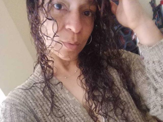 A close-up selfie of Angelonix. Angelonix has curly, wet brown hair falling over her face and shoulders. She's wearing a gray, ribbed, V-neck sweater with a slightly open neckline. Her light brown skin is visible, and she has small hoop earrings. Her right hand is raised, touching her hair. The image is slightly blurry, focusing on Angelonix's face and upper torso. Her expression is neutral, with slightly parted lips. The background is indistinct, with a patterned object partially visible on the right.