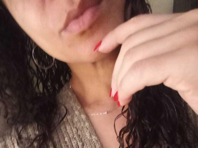 Close-up of Angelonix's face and hand. Angelonix has curly black hair, wears large silver hoop earrings, and a beige textured sweater. Her red manicured fingers gently touch her chin. Soft, warm lighting highlights her light brown skin. The image focuses on her slightly parted lips and the delicate silver necklace she wears. The composition emphasizes her facial features and hand.